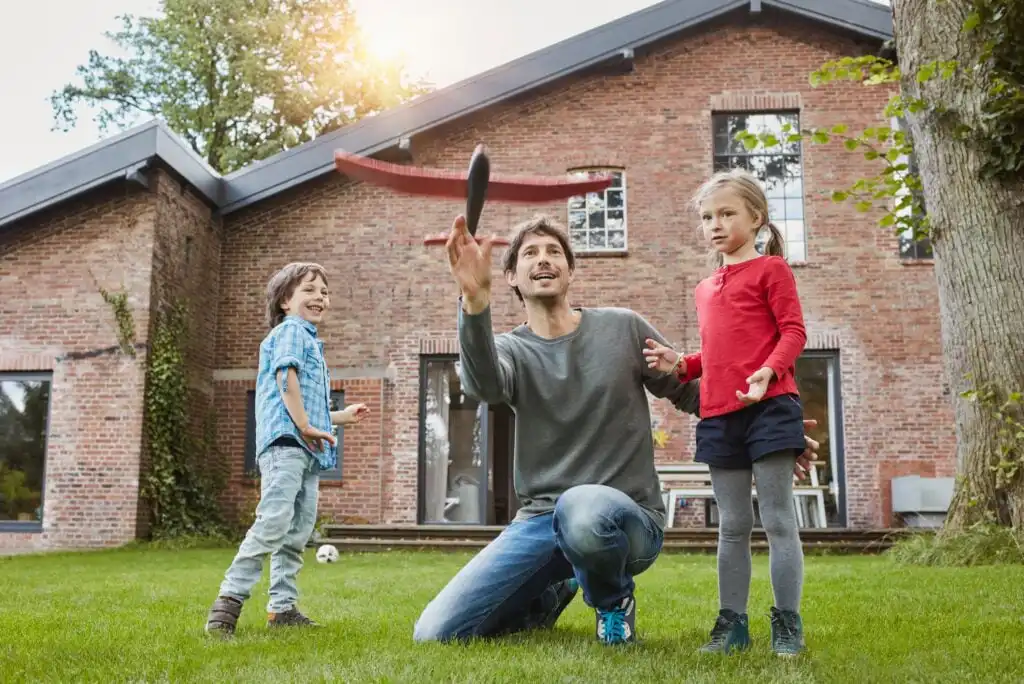 A man kneeling on grass tosses a frisbee while a smiling boy and girl stand nearby in front of a brick house, enjoying a sunny day outdoors.