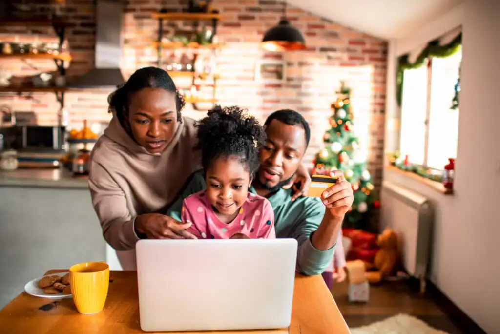 A family of three sits at a table using a laptop in a cozy kitchen decorated for Christmas. The parents assist their young daughter, and the father holds a credit card. A yellow mug and cookies are on the table.