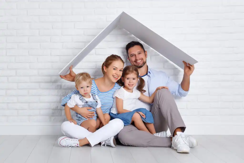 A smiling family of four sits on the floor against a white brick wall, holding a cardboard roof over their heads, symbolizing a home. The parents sit with their young son and daughter.