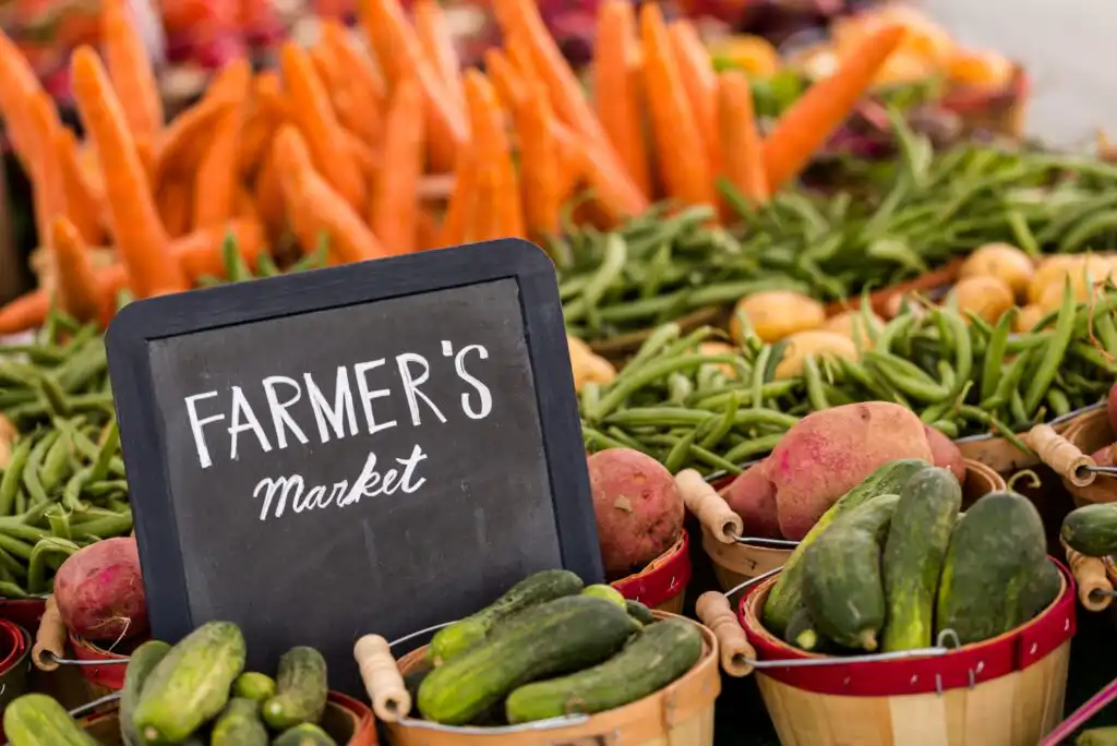 A chalkboard sign reading Farmers Market is displayed in front of baskets filled with fresh vegetables, including cucumbers, potatoes, green beans, and carrots. More produce is visible in the background.