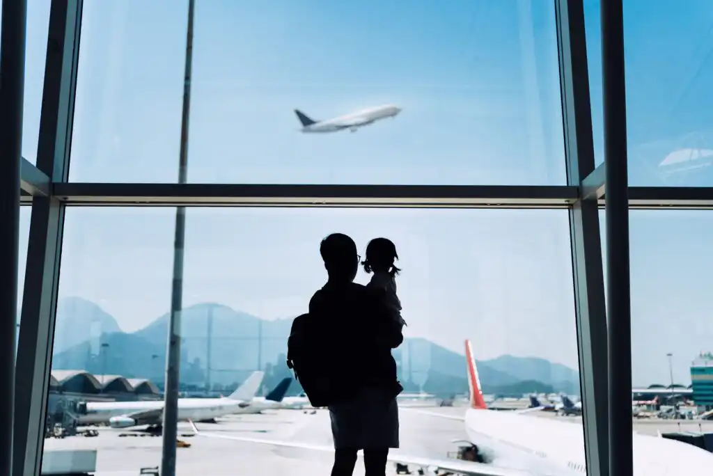 A silhouette of an adult holding a child while looking out an airport window at planes on the runway and one airplane flying in the sky, with mountains visible in the distance.