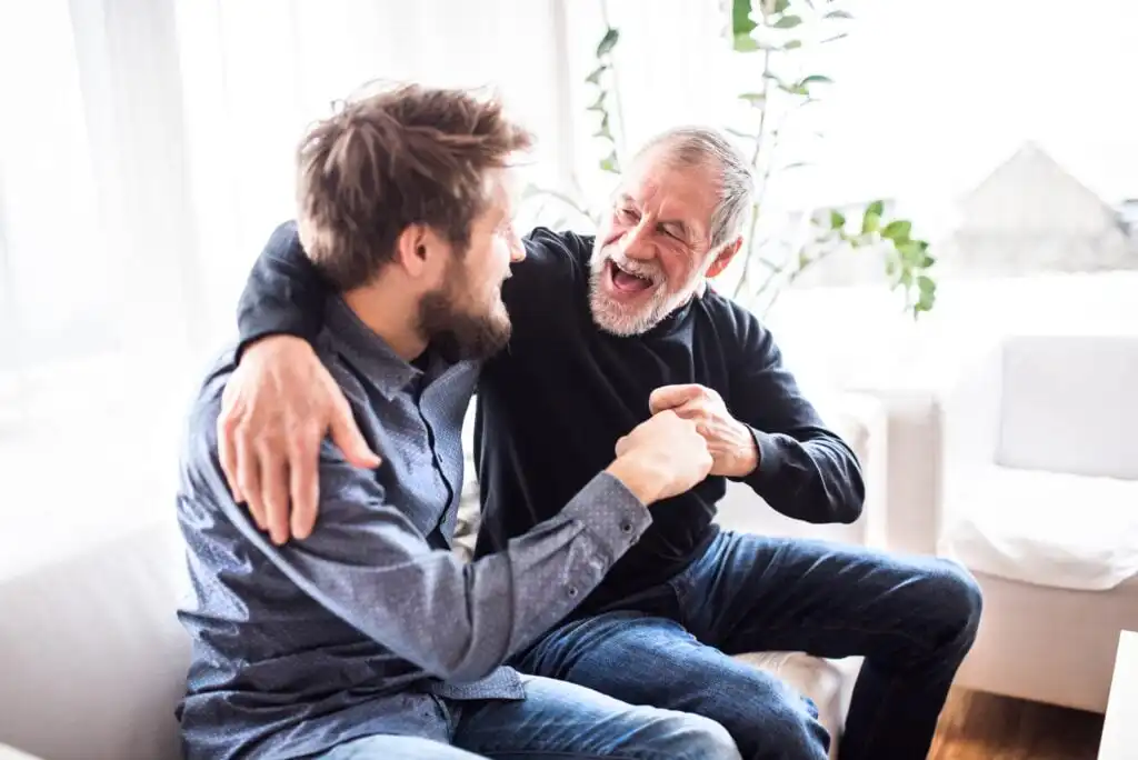 An older man and a younger man sit on a couch, laughing and hugging each other with joy. They appear to be sharing a happy, lighthearted moment in a bright living room.