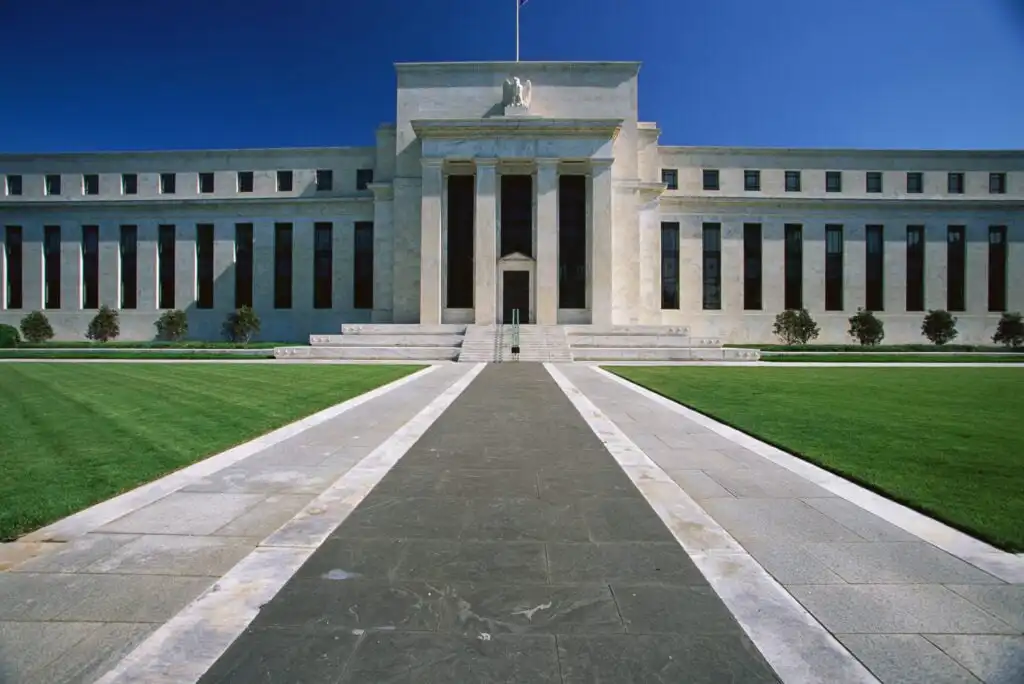 A wide walkway leads to a large, white, neoclassical government building with tall columns, symmetrical windows, and green lawns under a clear blue sky.