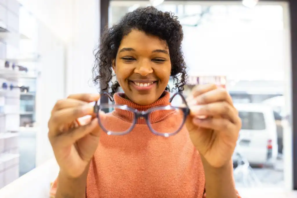 A smiling woman in a peach sweater holds a pair of eyeglasses in front of her, inside a bright eyewear store.