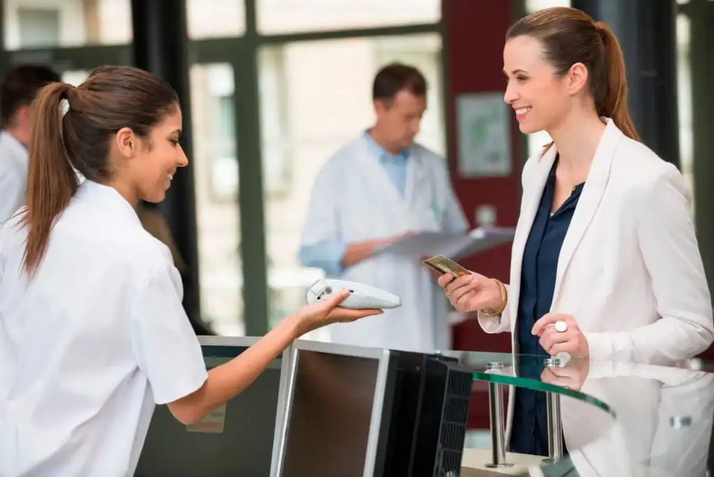 A woman in a white blazer smiles while handing her credit card to a receptionist in a medical office. Two people in lab coats are visible in the background, standing and talking.