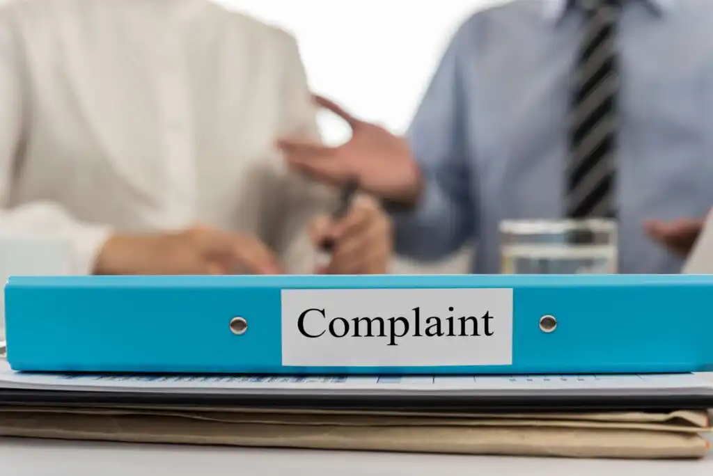 A blue binder labeled Complaint sits on a desk with papers. In the blurred background, two people are having a discussion, with one gesturing and the other holding a pen.