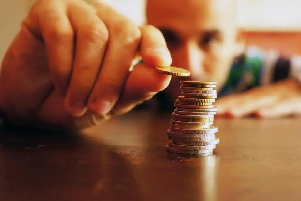 A person’s hand carefully stacks coins on a table, with several small piles of coins in focus and the person’s face blurred in the background.