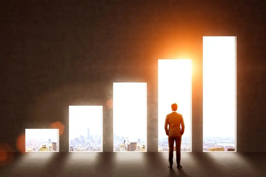A person in a suit stands facing a glowing bar graph with increasing bars, sunlight streaming through the tallest bar, against a cityscape background, symbolizing growth and progress.