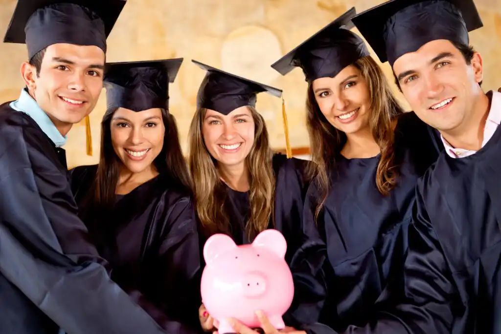 Five smiling graduates in caps and gowns stand close together, holding a pink piggy bank in front of them, suggesting themes of saving money, scholarships, or financial planning for education.