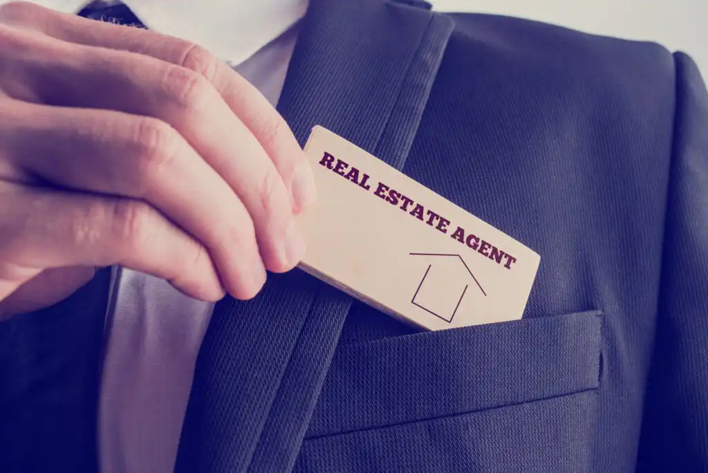A person in a suit puts a wooden card labeled REAL ESTATE AGENT with a house icon into their jacket pocket.