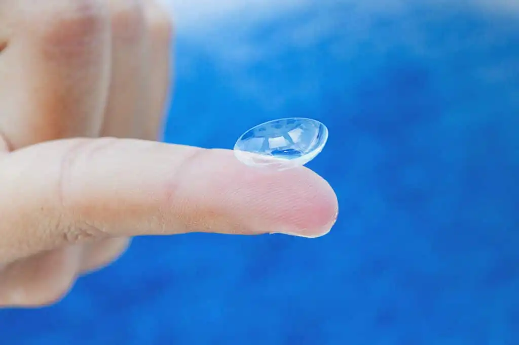 A close-up of a hand holding a clear contact lens on the tip of an index finger with a blurred blue background.
