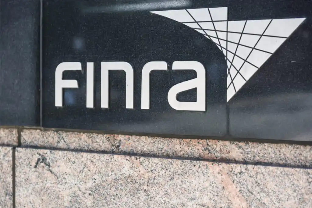 The image shows the FINRA logo displayed on a black and stone wall, featuring white block letters and a geometric design to the right of the text.