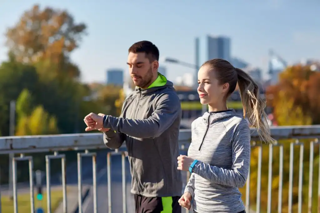 A man and a woman wearing athletic clothing are jogging outdoors on a bridge. The man is checking his watch, while the woman is smiling. Trees and city buildings are visible in the background.