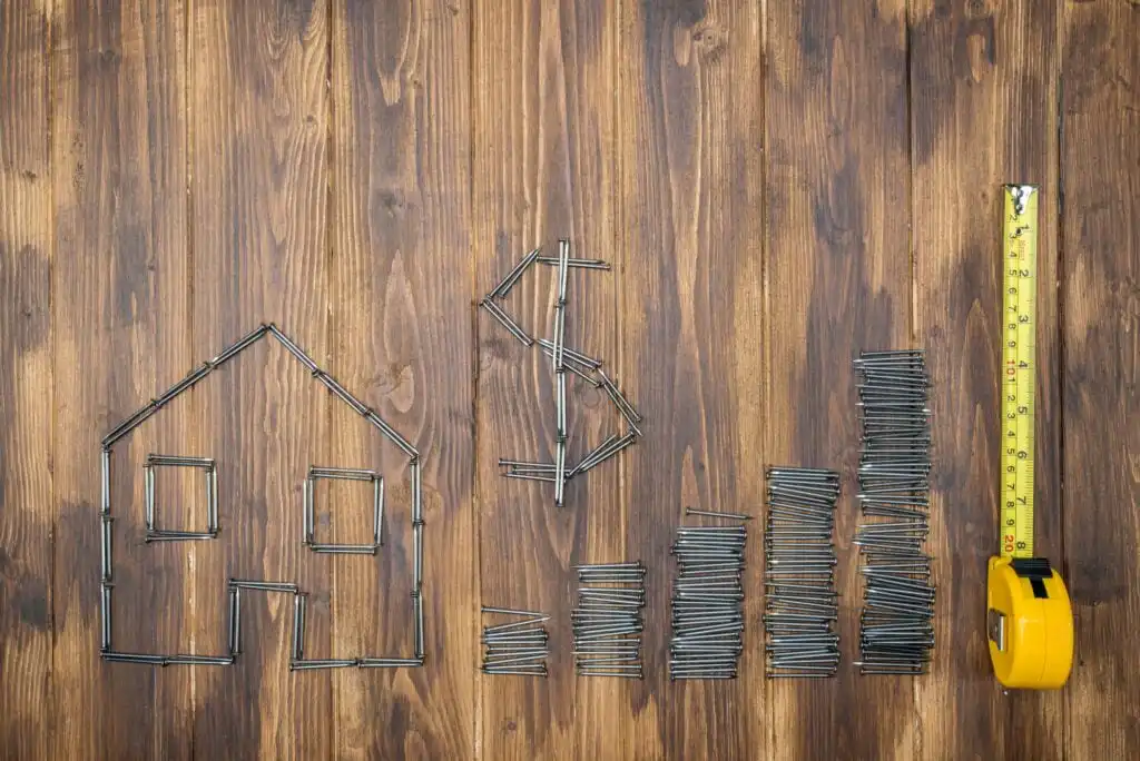 Nails arranged on a wooden surface form the shape of a house, a dollar sign, and ascending bar charts. A yellow measuring tape lies vertically on the right side.