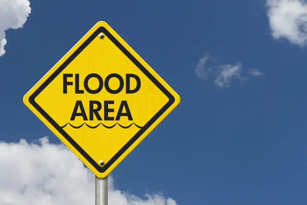 A yellow diamond-shaped road sign with the words FLOOD AREA and wavy lines, indicating possible flooding. The sign is set against a blue sky with some white clouds.