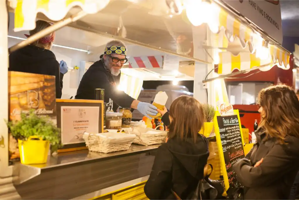 A man wearing a hat and glasses serves food from a brightly lit food truck to two women standing outside. The scene is lively and cheerful, with yellow decorations and baskets visible on the counter.