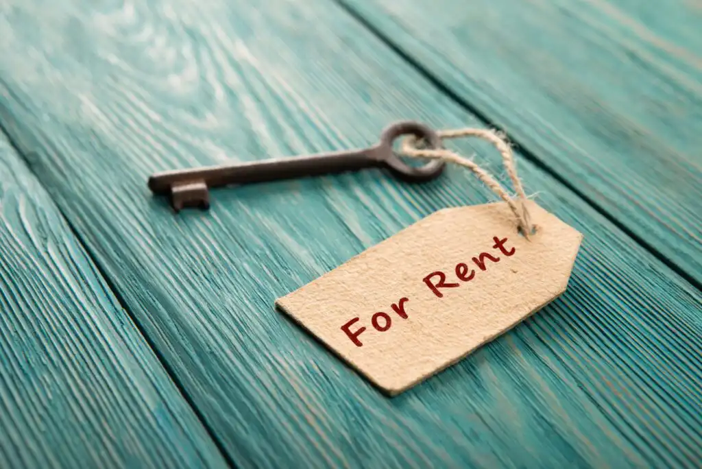 An old-fashioned key lies on a teal wooden surface with a brown paper tag attached by string. The tag has For Rent written on it in red letters.