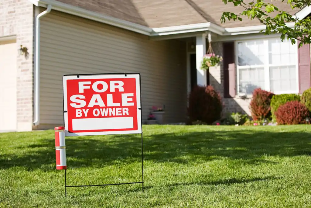A For Sale By Owner sign is staked in the front yard of a suburban house with beige siding, a porch, and green grass, suggesting the house is being sold directly by the owner.