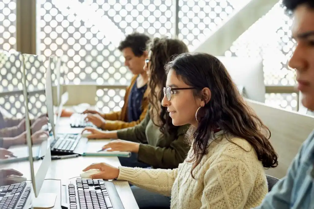 Several young adults sit in a row at computer desks, focused on their monitors. The background features a wall with circular patterns letting in natural light.