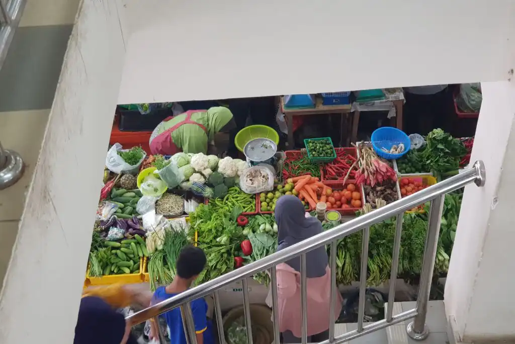 A view from above of people buying and selling assorted fresh vegetables, including carrots, lettuce, cauliflower, and tomatoes, at a market stall. The scene is partially framed by stair railings.