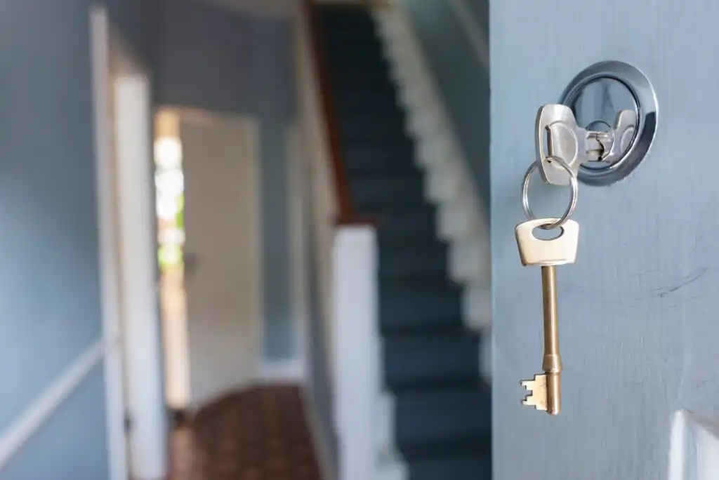 A close-up of a key inserted in a door lock, with a staircase and hallway blurred in the background, inside a home.
