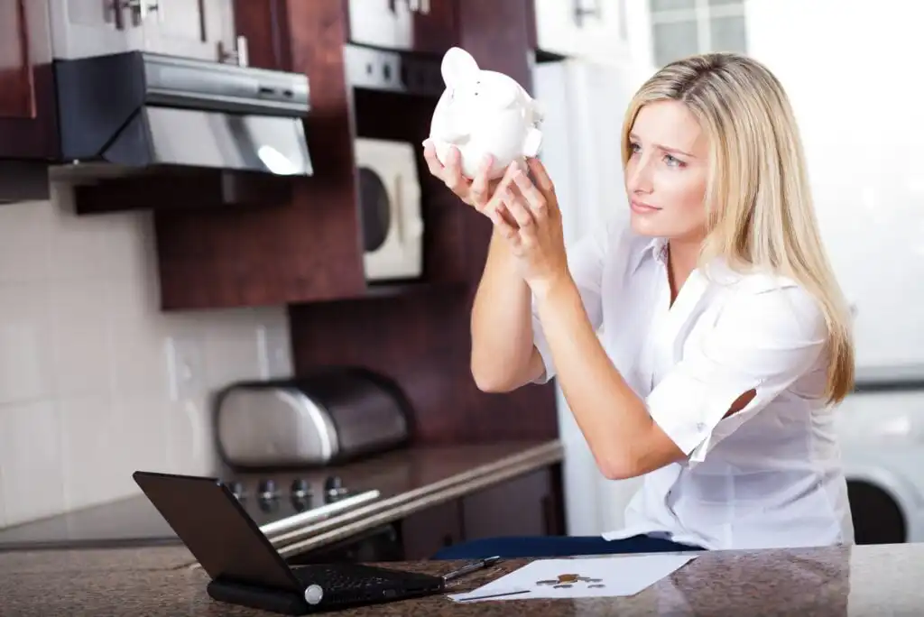 A woman sitting at a kitchen counter shakes a white piggy bank while looking concerned. There are coins and a piece of paper on the counter beside a laptop.