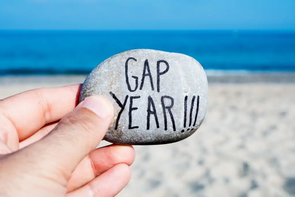 A hand holds a smooth stone with the words GAP YEAR!!! written on it, with a sandy beach and the blue sea in the background.