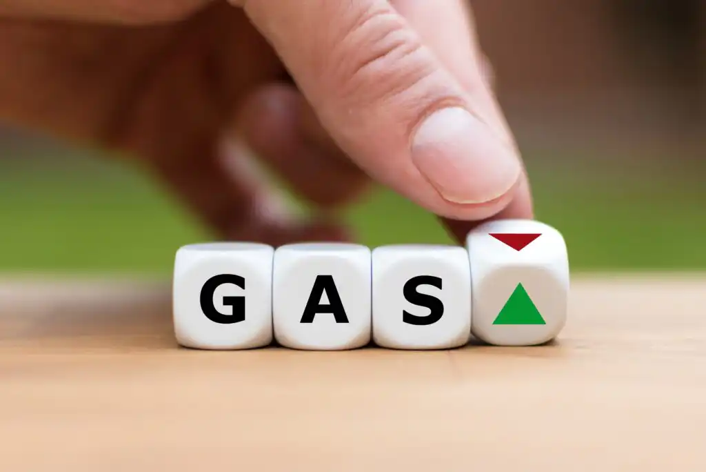 A hand flips a dice next to three others spelling GAS. The flipped dice shows a green upward triangle and a red downward triangle, symbolizing rising or falling gas prices.