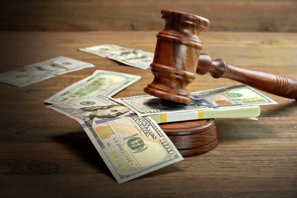A judge’s wooden gavel rests on a stack of US hundred-dollar bills on a wooden table, with more scattered bills in the background.