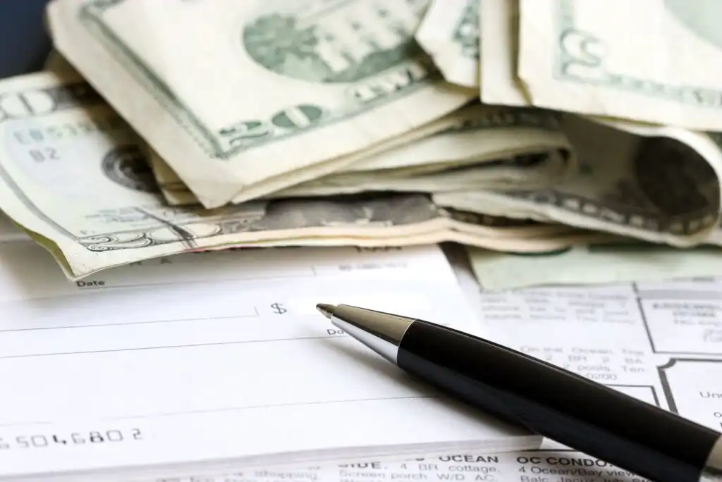 A close-up of a blank check, a pen, and a stack of U.S. dollar bills placed on top of financial documents.