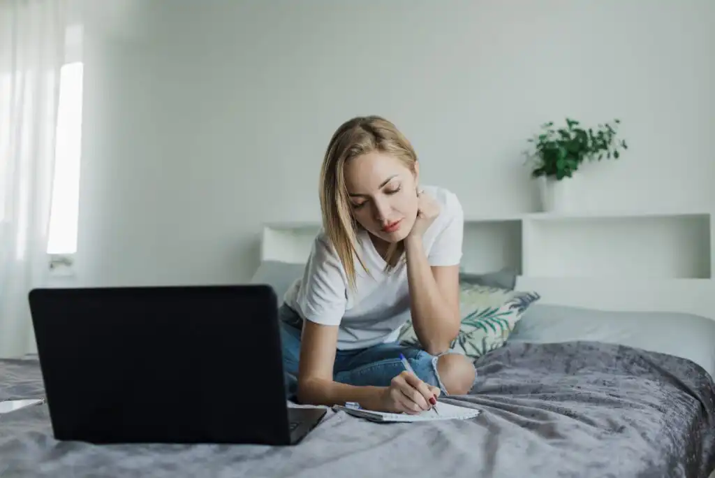 A woman sits on a bed, leaning over a notebook and writing, with a laptop open in front of her. She appears focused. The room is bright and modern, with a plant and cushions in the background.
