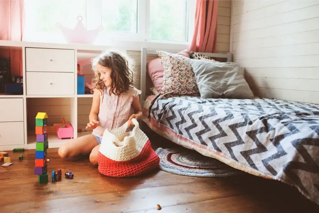 A young girl sits on the floor in a sunlit bedroom, placing toys into a red and white basket. Colorful blocks are stacked beside her, and the bed with patterned bedding is nearby. The room has wood floors and a window with pink curtains.