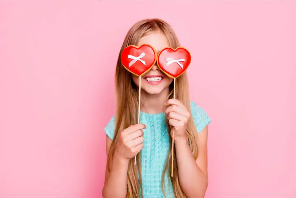 A smiling girl with long blonde hair holds two heart-shaped cookies on sticks over her eyes, standing against a pink background. She wears a light blue dress.