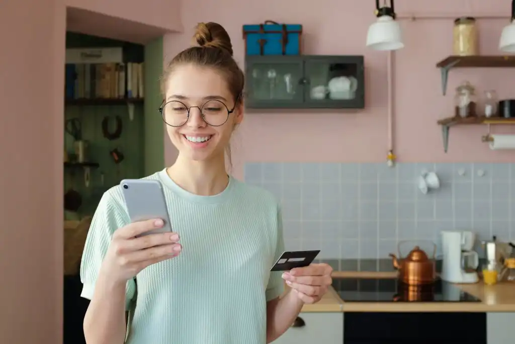 A young woman with glasses stands in a kitchen, smiling at her smartphone while holding a credit card. The kitchen has light blue tiles, shelves, and various kitchenware in the background.