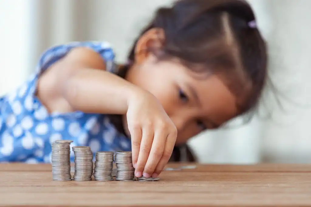 A young girl in a blue polka dot dress carefully stacks coins into piles on a wooden table, focusing intently on her task.