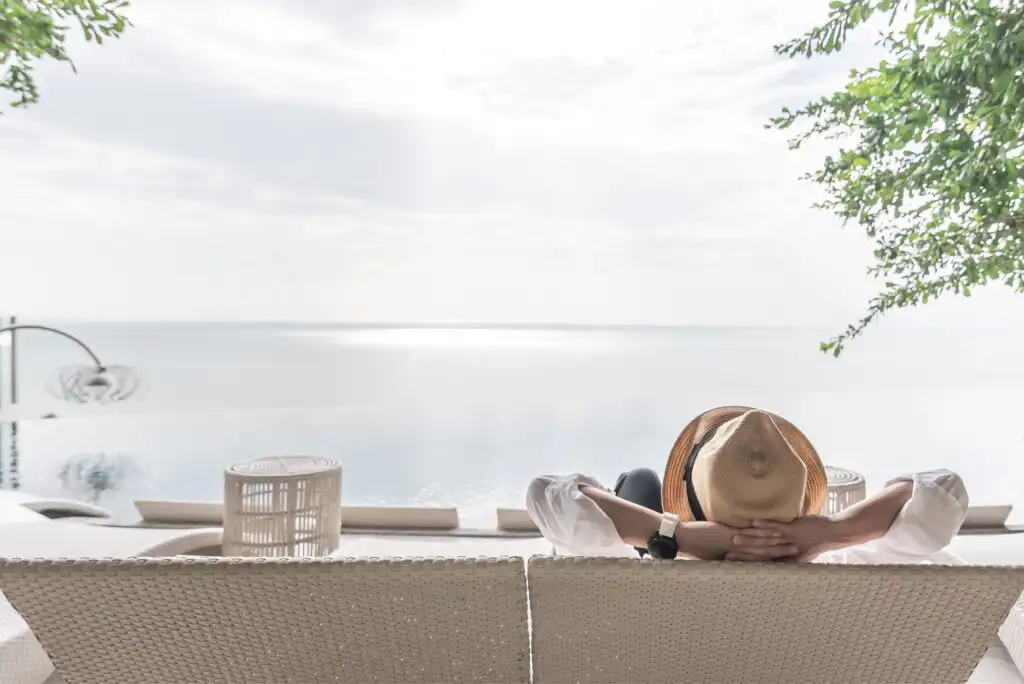 Person wearing a straw hat and white shirt relaxes on a lounge chair, facing a calm infinity pool and expansive sea under a bright, cloudy sky, with greenery overhead on the right.