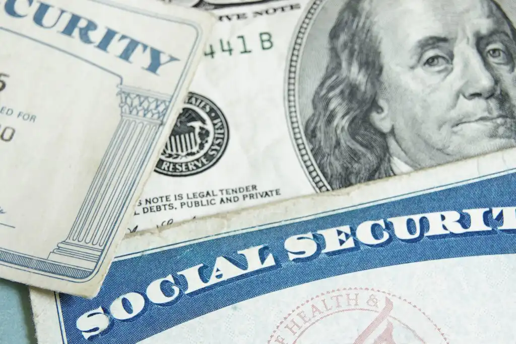 Close-up of two Social Security cards and a U.S. one hundred dollar bill, partially overlapping, symbolizing finances and Social Security benefits.