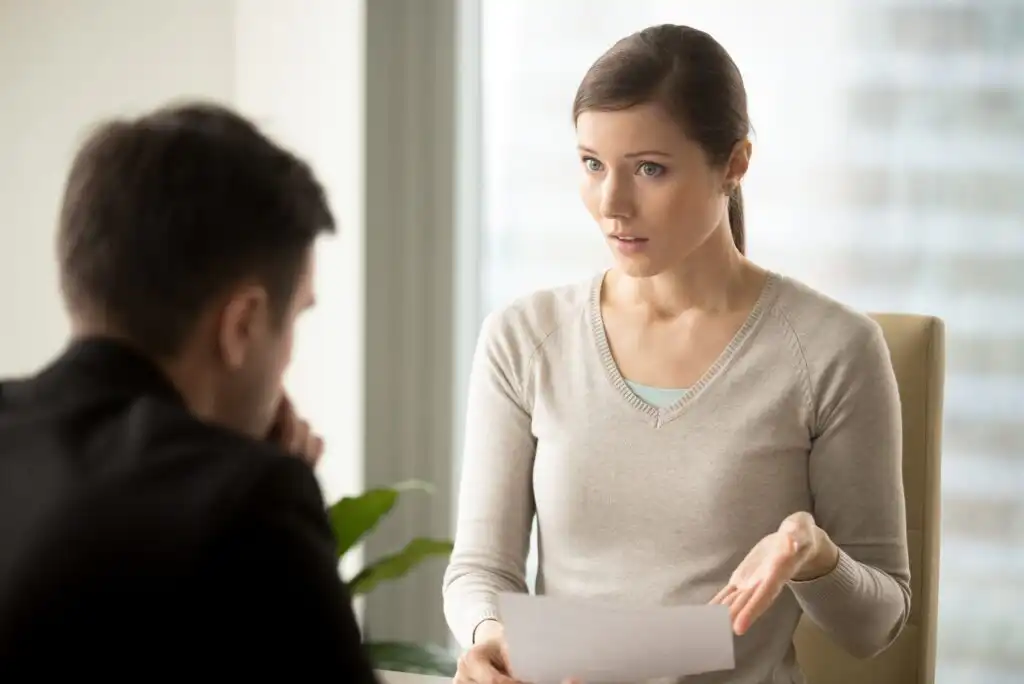A woman holding a document speaks with a concerned expression to a seated man in an office setting. Light comes through a large window behind them.