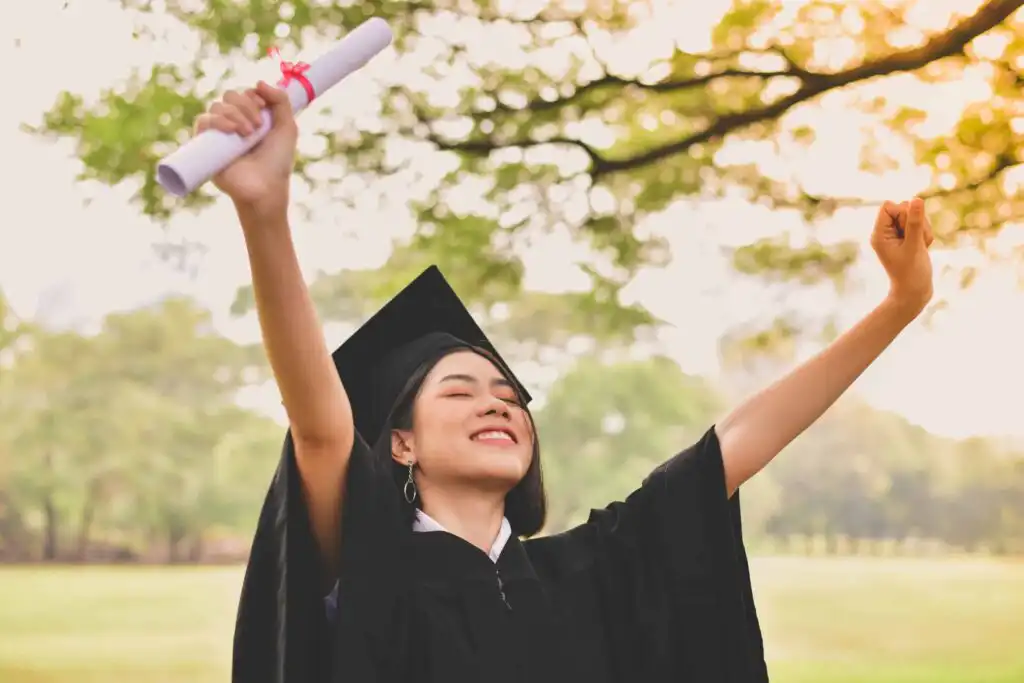 A young woman in a graduation cap and gown stands outdoors, smiling joyfully with her arms raised. She holds a diploma in one hand, celebrating her achievement with trees and sunlight in the background.