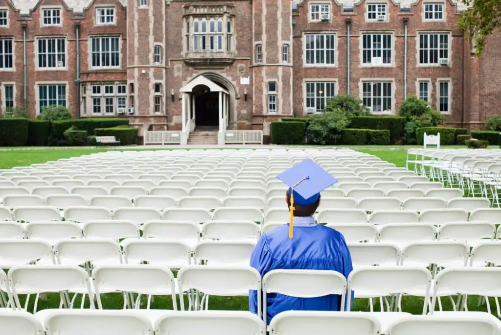 A person in a blue graduation gown and cap sits alone among rows of empty white chairs facing a large brick school building, suggesting a solitary graduation ceremony.