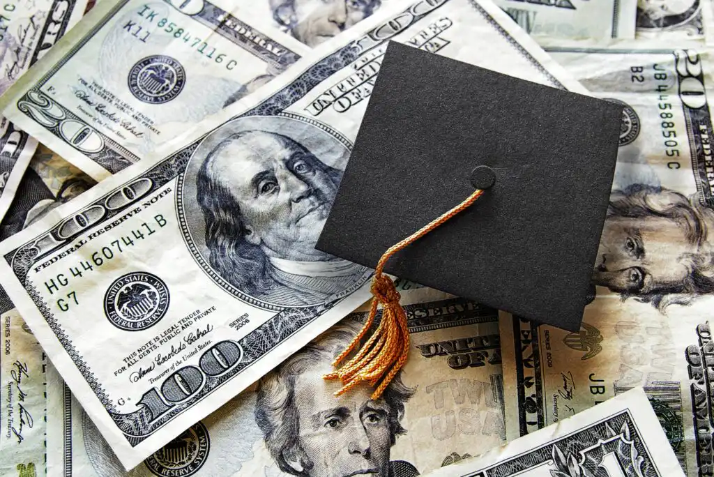 A black graduation cap with a gold tassel rests on top of scattered U.S. dollar bills, symbolizing the cost of education or student loans.