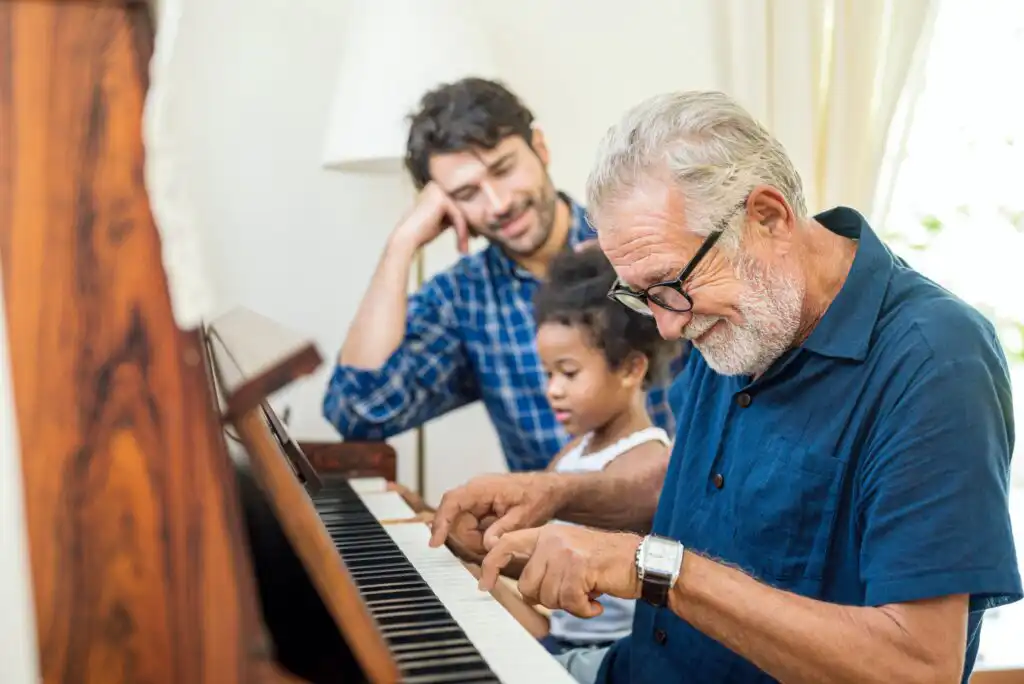 An elderly man plays piano with a young girl, while a man in a checkered shirt looks on, smiling. All three appear to be enjoying the moment in a cozy, well-lit room.