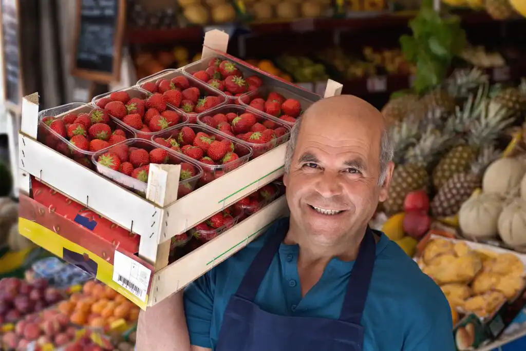 A smiling vendor in a blue shirt and apron holds boxes of fresh strawberries on his shoulder at a colorful fruit market, with various fruits displayed in the background.