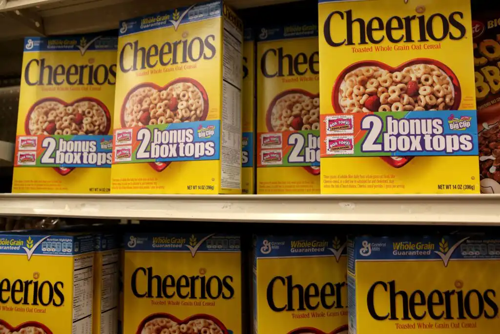 Several yellow Cheerios cereal boxes are displayed on supermarket shelves, with 2 bonus box tops labels visible on the front of each box. The boxes show a bowl filled with cereal and milk.