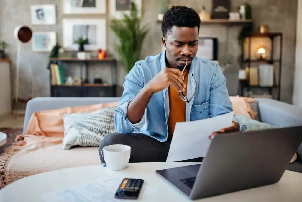 A man sits on a sofa at home, holding eyeglasses and reading a document. A laptop, coffee mug, and papers are on the table in front of him. He appears focused, with modern decor in the background.