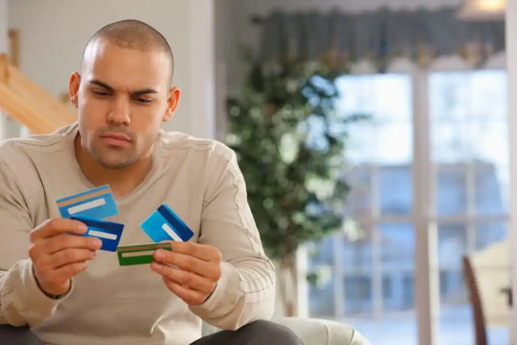 A man sits indoors, looking thoughtfully at four credit cards in his hands. The background shows a bright room with a large window, some greenery, and neutral-colored furnishings.