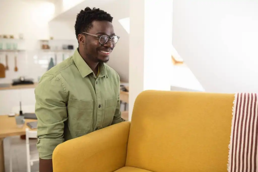 A man wearing glasses and a green shirt smiles while moving a yellow sofa in a bright, modern living room with a red-and-white striped blanket on the armrest.