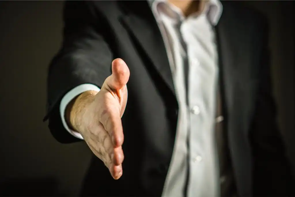 A person wearing a black suit and white shirt extends their right hand forward, as if offering a handshake, with the background blurred and out of focus.