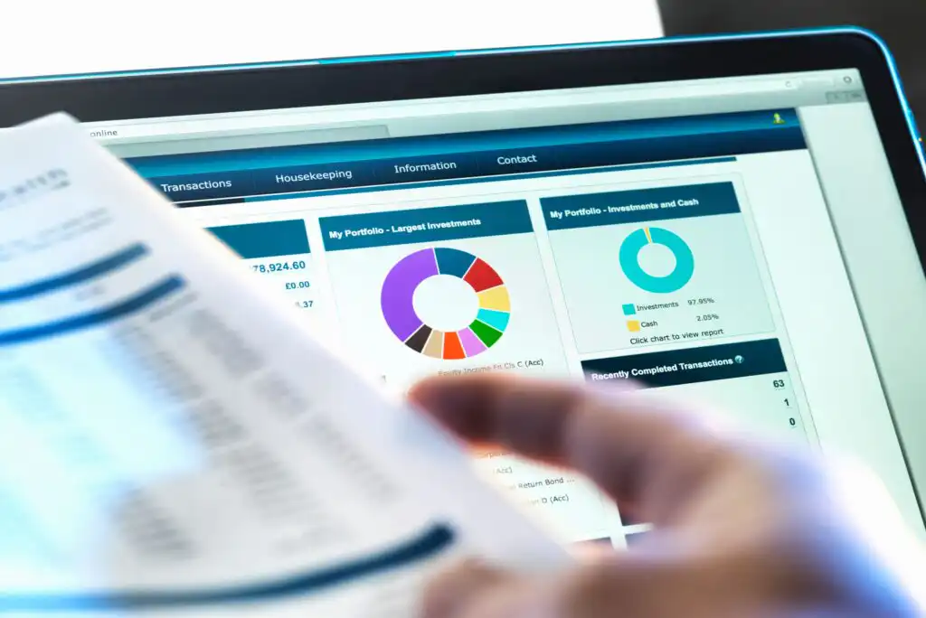 A person reviews financial documents in front of a computer screen displaying investment portfolio charts, including a pie chart and bar graphs, with blue and white interface elements.