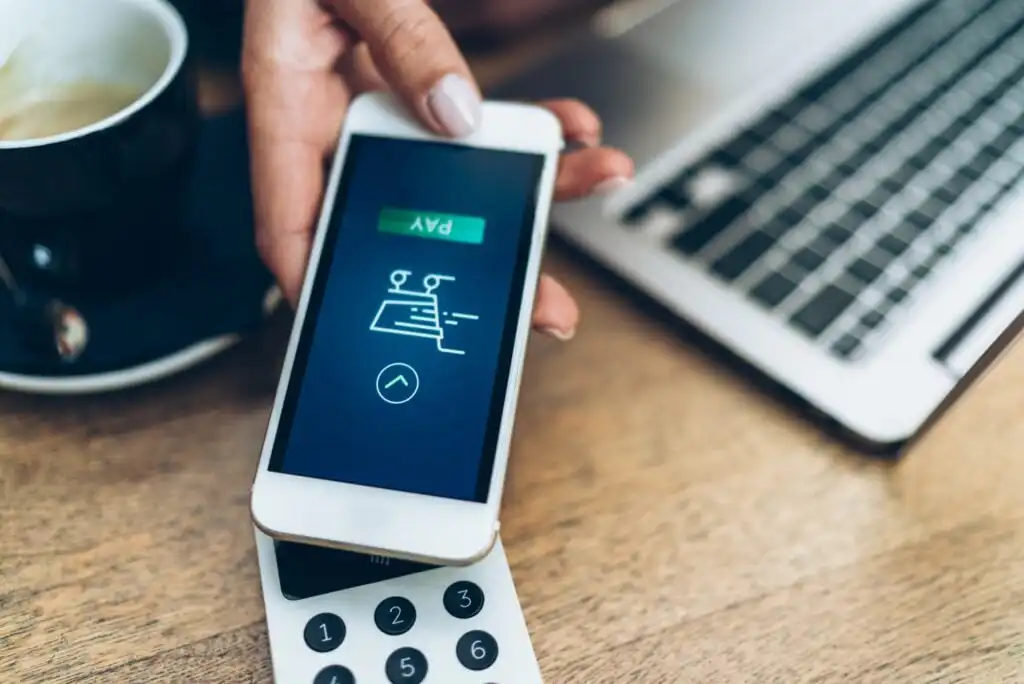 A person holds a smartphone displaying a Pay screen over a card reader, making a contactless payment, with a laptop and coffee cup in the background.